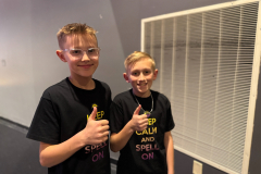 Two boys giving thumbs up in front of a ventilation grate.