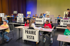 Students seated at desks in a classroom setting, participating in a spelling bee with numbers and names displayed on signs