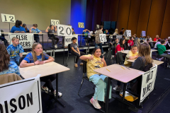 Students seated at desks in a classroom setting, participating in a spelling bee competition.