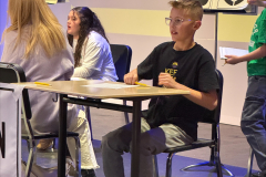 This is a black and white photograph of a young boy sitting at a table in front of a sign that reads "2025 Elementary Spell