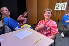 In a classroom setting, a group of people are gathered around tables, engaged in an activity. A young boy is seated at a