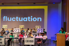 Children participating in a spelling competition, standing at desks with numbers on them, indicating their positions. The