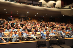 Auditorium filled with people at an event, sitting on chairs in rows facing a stage where a group of children wearing blue