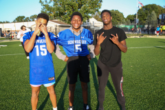 Three young men standing on a soccer field, posing for a picture. They are wearing blue and white jerseys, and one of them