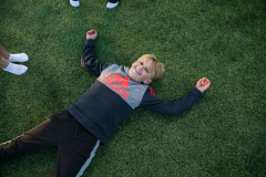 Young boy lying on grassy field, arms spread out, smiling broadly.