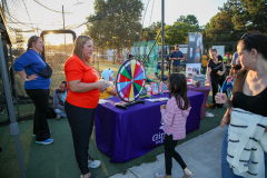 Woman in red shirt stands near wheel of fortune at outdoor event, surrounded by people.