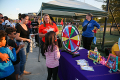 People gathered around a table with a wheel of fortune and prizes at an outdoor event.