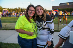 Two women posing together at a sports event, one wearing a cheerleader uniform, both smiling and looking towards the camera.