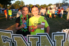 Two people standing in front of a sign at an event. The sign says "GNTN". One person is holding a drink and smiling towards