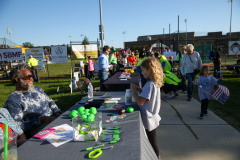 People gathered around a table at an outdoor event, with some standing and others seated on chairs, engaging in conversation.