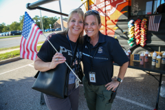 Two women posing at a food truck event. One woman is holding an American flag. The other woman is wearing a badge and has a