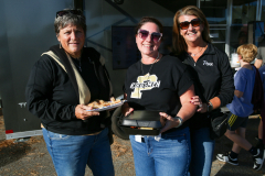 Three women posing together at an event, smiling and holding food items, standing in front of a trailer with a sign that