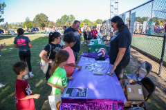 People gathered around a table at an outdoor event, possibly a fundraiser or fair, with various items on display and a woman