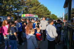 People gathered outside a food truck at an event, enjoying their meal in a park-like setting on what appears to be a sunny