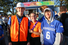 Three people posing at an event, two in orange vests and one in a blue jersey, standing in front of a food truck.