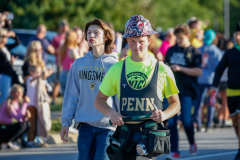 Two people walking together at a parade. One person is wearing a hat and the other has a sign around their neck.