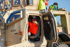 Man in red shirt sitting inside military truck at parade.