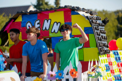 Two men in colorful shirts standing on a float decorated to look like a house, celebrating an event.
