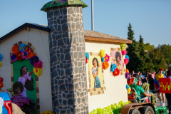Colorful display of a float in a parade, featuring a building decorated with various signs and images, including a woman's