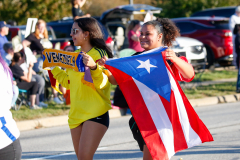 Two women holding flags, one with a yellow shirt and the other in a black shirt, walking together in a parade.