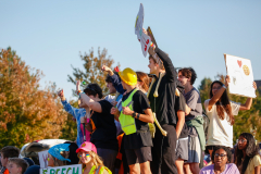 Group of people standing on a stage at a parade, holding signs and waving to the crowd.