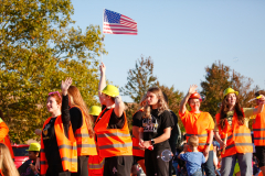 Group of people wearing safety vests and hard hats at a parade, some holding signs and waving, in front of a flag.