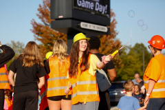 People in safety vests standing by a sign, blowing bubbles, with construction equipment in the background.