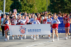 Cheerleaders marching in a parade, holding banner with logo and text.