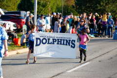 People gathered at a parade, walking down a street carrying signs and banners, including one that says 'Dolphins'.