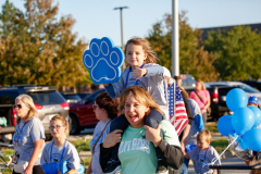 Woman carrying child on shoulders at parade, both wearing blue dog ears.