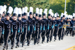 A group of marching band members in uniforms, standing in a line on a street.
