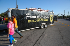 People walking past a black trailer advertising Penn High School's building trades program during a parade.
