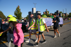 People walking in a parade, some holding signs, all wearing safety vests.