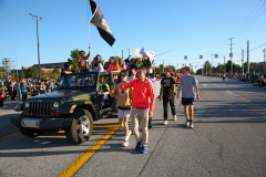 People walking down a street in a parade, with a jeep behind them. The scene is set during the daytime under clear skies.