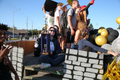 People celebrating at a parade, standing on a stage made of cardboard bricks and balloons, holding trophies and signs.