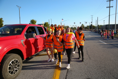 Group of people in orange vests walking down street, leading parade float.