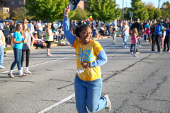 Woman in yellow shirt running through parking lot during parade.