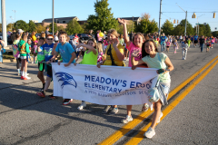 Group of people posing for a picture at a parade, holding a banner that reads "Meadow's Elementary School".
