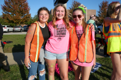 Three young women wearing orange safety vests standing in front of a building. They are smiling and posing for the camera.