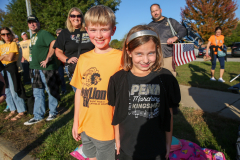 Two children posing in front of a crowd at a parade, both wearing matching shirts and standing on a sidewalk.