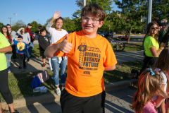 Boy in orange shirt giving thumbs up at parade.