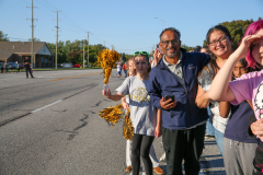 A group of people gathered on a street corner during a parade, holding balloons and flags, smiling at the camera.