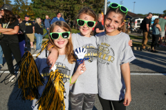 Three young girls posing for a picture at a parade, wearing festive shirts and sunglasses, standing in front of a crowd of
