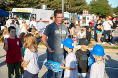 Man in a black shirt standing next to children in blue shirts holding baseball bats.