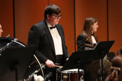 Three musicians performing on stage. One person playing a drum set, another with a saxophone, and one holding a microphone.