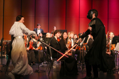 Orchestra performing in a theater, two people dressed as Darth Vader and Princess Leia from Star Wars, conductor at podium.
