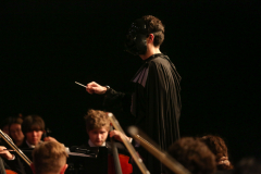 A conductor in a black cape stands on stage directing an orchestra, holding a baton and gesturing with his hands.