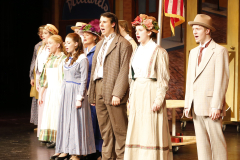 Group of actors on stage dressed in period costumes, performing in a musical production.