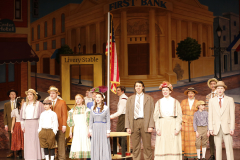 Theatrical production of "The Music Man" on stage with a large cast in costumes, performing on a set that resembles an old