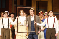 Group of people on stage in a theater, dressed in costumes from the musical 'Oklahoma!', posing for a picture with a frame.